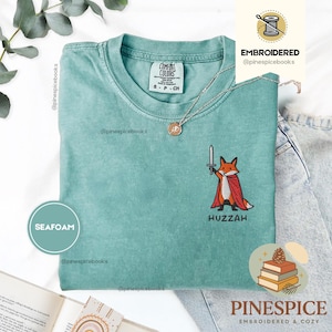 Peut inclure: Un t-shirt à manches courtes vert mousse avec une broderie représentant un renard portant une cape rouge et tenant une épée, accompagné du texte HUZZAH. Le vêtement présente une encolure ronde et une étiquette Comfort Colors au col.