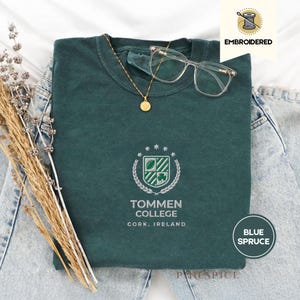 Pode incluir: Uma camiseta verde-azulado escuro com um brasão bordado com um escudo, trevo e estrelas. O texto "TOMMEN COLLEGE CORK, IRELAND" está abaixo. Um colar de ouro e óculos transparentes repousam sobre a camisa.