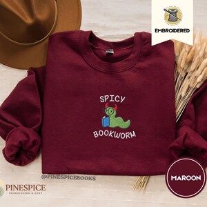 Puede incluir: Sudadera burdeos de cuello redondo con el texto bordado "SPICY BOOKWORM" y un personaje de gusano verde con gafas y un libro. La sudadera tiene mangas largas y cuello redondo. La marca es "PINESPICE".