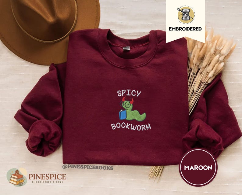 Puede incluir: Sudadera burdeos de cuello redondo con el texto bordado "SPICY BOOKWORM" y un personaje de gusano verde con gafas y un libro. La sudadera tiene mangas largas y cuello redondo. La marca es "PINESPICE".