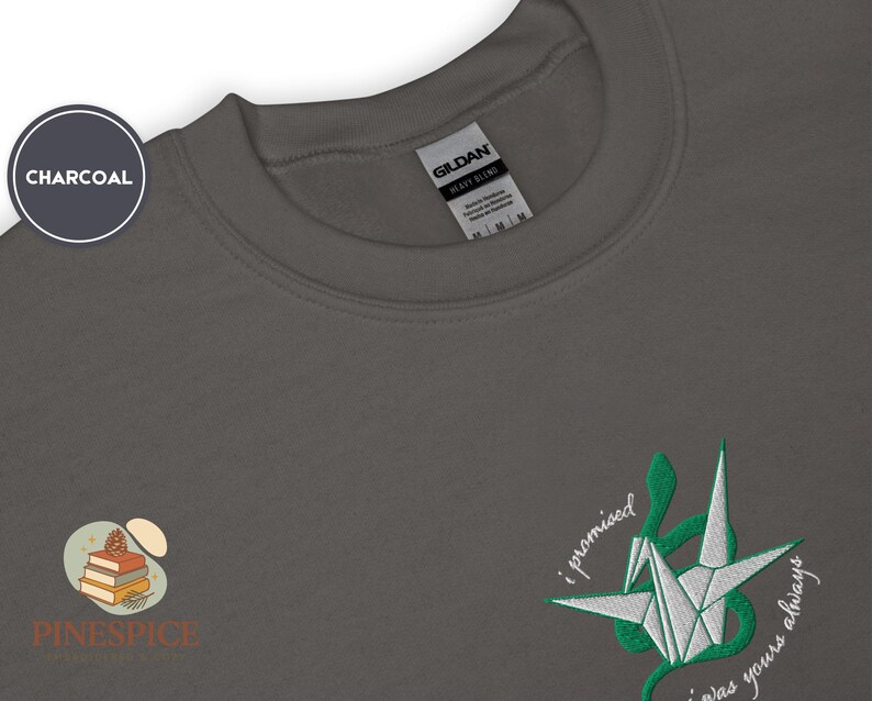 K&ouml;nnte beinhalten: Anthrazitfarbenes Sweatshirt mit Rundhalsausschnitt. Es hat ein gesticktes Origami-Kranich-Design in Wei&szlig; und Gr&uuml;n mit dem Text "i promised i was yours always". Das Markenlogo "Pinespice" ist ebenfalls sichtbar.