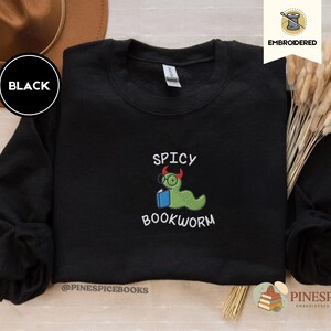 Puede incluir: Sudadera negra bordada con el texto "Spicy Bookworm" y un gusano de libro de dibujos animados con gafas y un libro. La sudadera tiene mangas largas y cuello redondo. La marca "Pinespice" est&aacute; en la parte inferior.