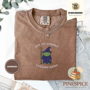 May include: Espresso-coloured t-shirt with the text "OFF TO COMMIT SHENANIGANS" and an embroidered cartoon frog wearing a wizard hat and robe. The shirt has a crew neck and short sleeves. A silver necklace is draped across the shirt.