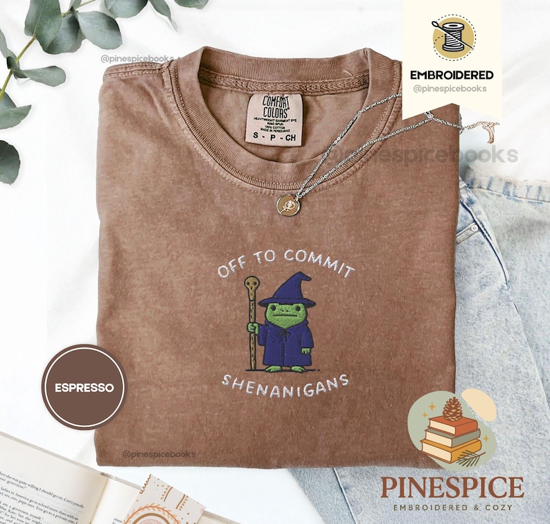 May include: Espresso-coloured t-shirt with the text "OFF TO COMMIT SHENANIGANS" and an embroidered cartoon frog wearing a wizard hat and robe. The shirt has a crew neck and short sleeves. A silver necklace is draped across the shirt.