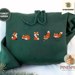 Könnte beinhalten: Ein waldgrüner Kapuzenpullover mit einer Stickerei von fünf kleinen orange-weißen Füchsen in verschiedenen Posen. Der Hoodie verfügt über passende grüne Kordelzüge am Ausschnitt sowie Textetiketten, die die Farbe und den Stickstil angeben.