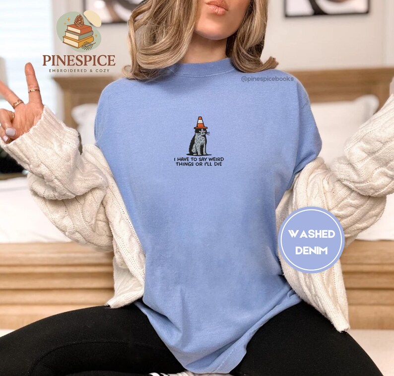 May include: A light blue washed denim sweatshirt with the text "I HAVE TO SAY WEIRD THINGS OR I'LL DIE". The sweatshirt features a graphic of a cat wearing a traffic cone hat. The brand name "PINESPICE" is at the top.