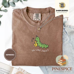 May include: A brown, crew-neck t-shirt with short sleeves. The shirt displays an embroidered green caterpillar playing a yellow saxophone, with the text "YA LIKE JAZZ?". The brand name "COMFORT COLORS" and size details are also visible.