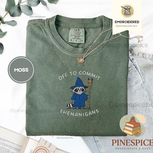 May include: A moss green t-shirt featuring the embroidered phrase "OFF TO COMMIT SHENANIGANS" and a raccoon in a wizard outfit. The shirt is a Comfort Colors brand, size S/P/CH.