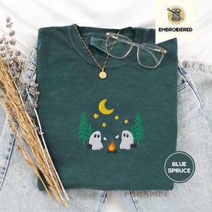 May include: A dark teal embroidered sweatshirt featuring a campfire scene with two ghosts, trees, a moon, and stars. The text "EMBROIDERED" is in the upper right corner, and "BLUE SPRUCE" is in a circle in the lower right corner. A gold necklace and glasses are on top.