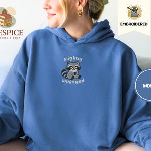 Pu&ograve; includere: Felpa con cappuccio blu indaco con un ricamo di un procione. Il procione &egrave; grigio e bianco con le parole "slightly unhinged" ricamate sotto. La felpa ha un cappuccio con coulisse e una tasca a marsupio.