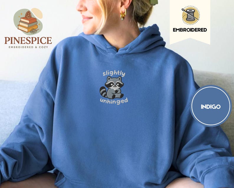 Pu&ograve; includere: Felpa con cappuccio blu indaco con un ricamo di un procione. Il procione &egrave; grigio e bianco con le parole "slightly unhinged" ricamate sotto. La felpa ha un cappuccio con coulisse e una tasca a marsupio.