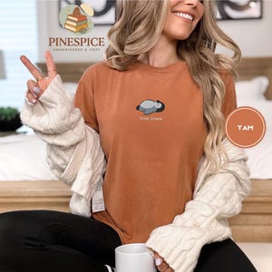 May include: A burnt orange t-shirt featuring the embroidered phrase "Diva Down" and a small penguin design. The shirt is from the Pinespice collection, embroidered and cozy.