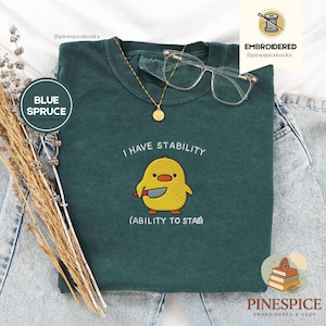 Könnte beinhalten: Blau-Fichte-T-Shirt mit dem gestickten Text "I HAVE STABILITY" und "(ABILITY TO STAB)". Das Shirt zeigt eine gelbe Küken-Grafik, die ein Messer hält. Eine goldene Halskette und eine klare Brille liegen auf dem Shirt.