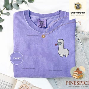 May include: A violet short-sleeved t-shirt featuring a small embroidered white llama holding a tiny red flower on the chest. The shirt has a crew neckline and a Comfort Colors brand label on the inside collar. The fabric has a washed-out appearance.