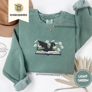 Op de afbeelding: Lichtgroene sweatshirt met een geborduurd ontwerp van een zwarte draak op een open boek, omringd door witte bloemen. De sweatshirt heeft lange mouwen en een ronde hals. De tekst "EMBROIDERED" staat in de linkerbovenhoek.