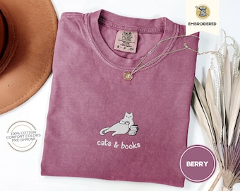 May include: Berry-colored Comfort Colors t-shirt with an embroidered cat design, holding a cup and resting on books. The text "cats & books" is below the cat. A gold necklace and a brown hat are also in the image.
