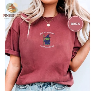 May include: A brick-red t-shirt featuring the phrase "OFF TO COMMIT SHENANIGANS" and a cartoon wizard. The wizard is green, wearing a blue robe and hat, and holding a staff. The shirt has a loose fit and is a casual style.
