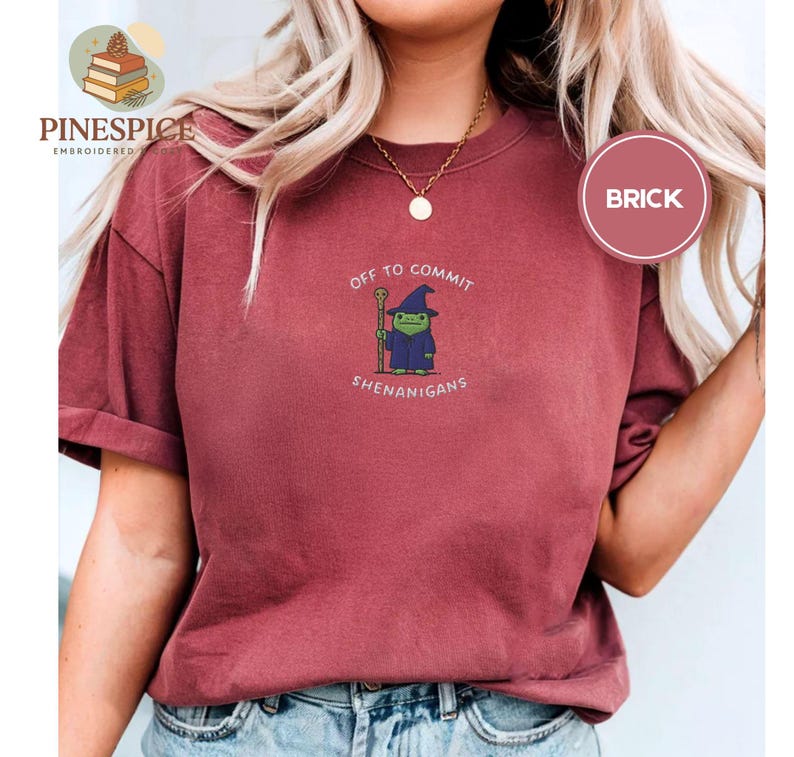 May include: A brick-red t-shirt featuring the phrase "OFF TO COMMIT SHENANIGANS" and a cartoon wizard. The wizard is green, wearing a blue robe and hat, and holding a staff. The shirt has a loose fit and is a casual style.