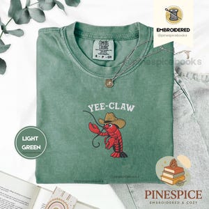 May include: Light green t-shirt with a crew neck and short sleeves. The shirt features an embroidered design of a red lobster wearing a cowboy hat, with the text "YEE-CLAW" above it. A silver necklace with a pendant is visible.
