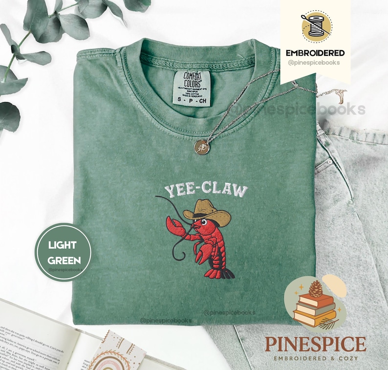 May include: Light green t-shirt with a crew neck and short sleeves. The shirt features an embroidered design of a red lobster wearing a cowboy hat, with the text "YEE-CLAW" above it. A silver necklace with a pendant is visible.