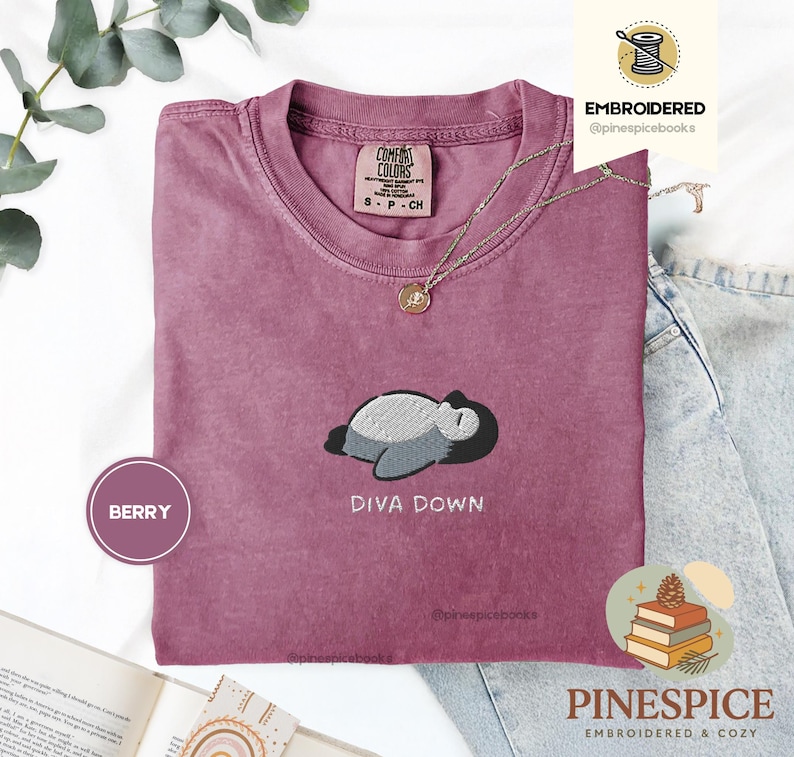 May include: A berry-coloured t-shirt featuring an embroidered design of a sleeping cartoon character and the text "DIVA DOWN." The shirt has a crew neck and short sleeves. A gold necklace with a pendant is draped on the shirt. The shirt is displayed on a white surface.