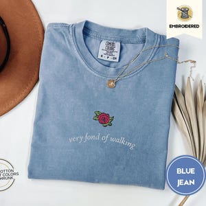 Puede incluir: Una camiseta azul vaquera Comfort Colors con una rosa bordada y la frase "very fond of walking". La camiseta tiene cuello redondo y mangas cortas. Un collar dorado y un sombrero marr&oacute;n tambi&eacute;n est&aacute;n en la imagen.