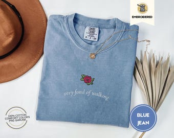 May include: A blue jean Comfort Colors t-shirt with an embroidered rose and the phrase "very fond of walking." The shirt has a crew neck and short sleeves. A gold necklace and a brown hat are also in the image.