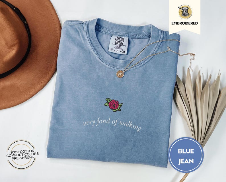 Puede incluir: Una camiseta azul vaquera Comfort Colors con una rosa bordada y la frase "very fond of walking". La camiseta tiene cuello redondo y mangas cortas. Un collar dorado y un sombrero marr&oacute;n tambi&eacute;n est&aacute;n en la imagen.