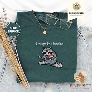 Puede incluir: Camiseta azul abeto con un mapache bordado sosteniendo libros y un cuchillo, con el texto "i require books". También se ven un collar dorado y gafas transparentes.