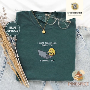 Op de afbeelding: Een blauwgroen t-shirt met de geborduurde tekst "I HOPE THIS EMAIL FINDS YOU BEFORE I DO" en een afbeelding van een kuiken met een laptop. Een gouden ketting en een heldere bril liggen op het shirt.