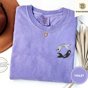 May include: A purple t-shirt with a black and white embroidered design of two fish. The shirt is made of 100% cotton and is pre-shrunk. The text "COMFORT COLORS" is visible on the tag.