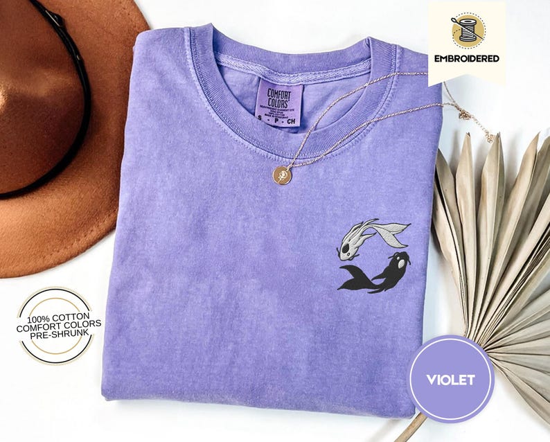 May include: A purple t-shirt with a black and white embroidered design of two fish. The shirt is made of 100% cotton and is pre-shrunk. The text "COMFORT COLORS" is visible on the tag.