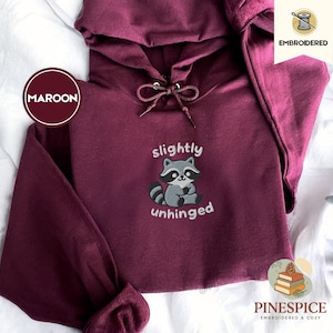 Pu&ograve; includere: Felpa con cappuccio bordeaux con cappuccio con coulisse e tasca a marsupio. La parte anteriore presenta una grafica ricamata di procione con le parole "slightly unhinged". La felpa con cappuccio &egrave; di Pinespice, ricamata e accogliente.