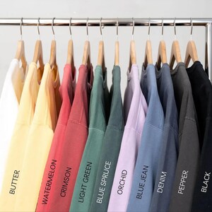 May include: Ten different colored sweatshirts hanging on a rack. The colors are white, butter, watermelon, crimson, light green, blue spruce, orchid, blue jean, denim, pepper, and black.