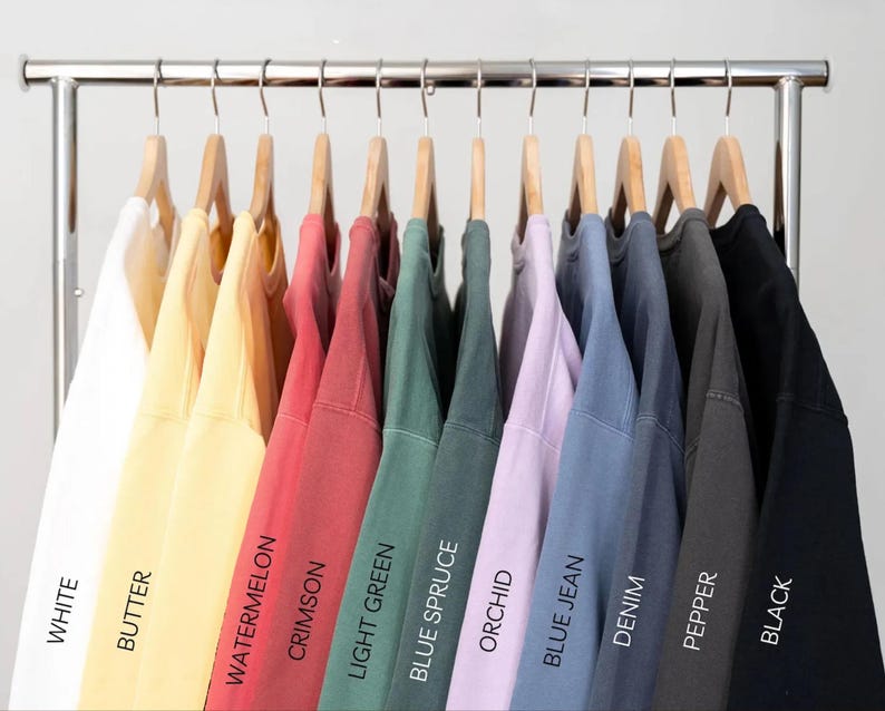 May include: Ten different colored sweatshirts hanging on a rack. The colors are white, butter, watermelon, crimson, light green, blue spruce, orchid, blue jean, denim, pepper, and black.