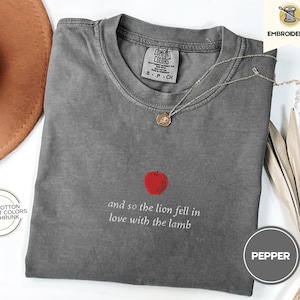 Peut inclure: Un t-shirt gris avec le texte "and so the lion fell in love with the lamb" et un graphique de pomme rouge. Le t-shirt est en 100% coton et pré-rétréci.