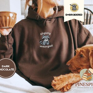 Pu&ograve; includere: Felpa con cappuccio marrone cioccolato scuro con la scritta ricamata "slightly unhinged" e una grafica di procione. La felpa ha un cappuccio con coulisse e una tasca a marsupio. Una tazza marrone &egrave; tenuta nella mano sinistra. Parte della collezione Embroidered & Cozy.