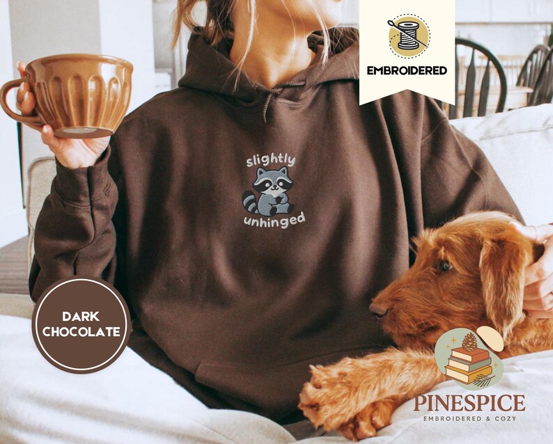 Pu&ograve; includere: Felpa con cappuccio marrone cioccolato scuro con la scritta ricamata "slightly unhinged" e una grafica di procione. La felpa ha un cappuccio con coulisse e una tasca a marsupio. Una tazza marrone &egrave; tenuta nella mano sinistra. Parte della collezione Embroidered & Cozy.