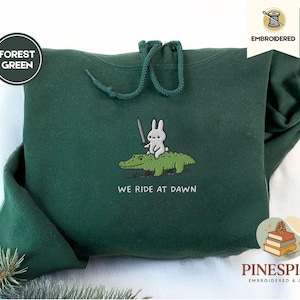 May include: Dark green hoodie with a drawstring. The front displays an embroidered design of a rabbit with a sword riding an alligator, with the text "WE RIDE AT DAWN". The hoodie is from Pinespice.