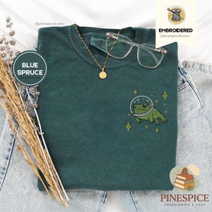 Puede incluir: Camiseta azul abeto con un diseño bordado de una rana con casco espacial. La camiseta también presenta un collar dorado, gafas transparentes y el texto "EMBROIDERED @pinespicebooks".