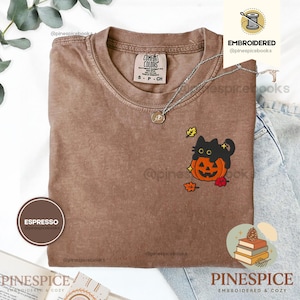 Puede incluir: Una camiseta color espresso con un diseño bordado de un gato negro sentado sobre una calabaza naranja de Halloween, rodeado de pequeñas hojas de otoño amarillas y rojas. La camiseta tiene cuello redondo y una etiqueta de la marca Comfort Colors.