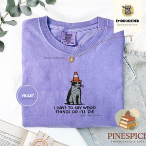 May include: A violet Comfort Colors t-shirt with an embroidered design of a cat wearing a traffic cone hat. The text "I HAVE TO SAY WEIRD THINGS OR I'LL DIE" is below the cat. A gold necklace is on the shirt.