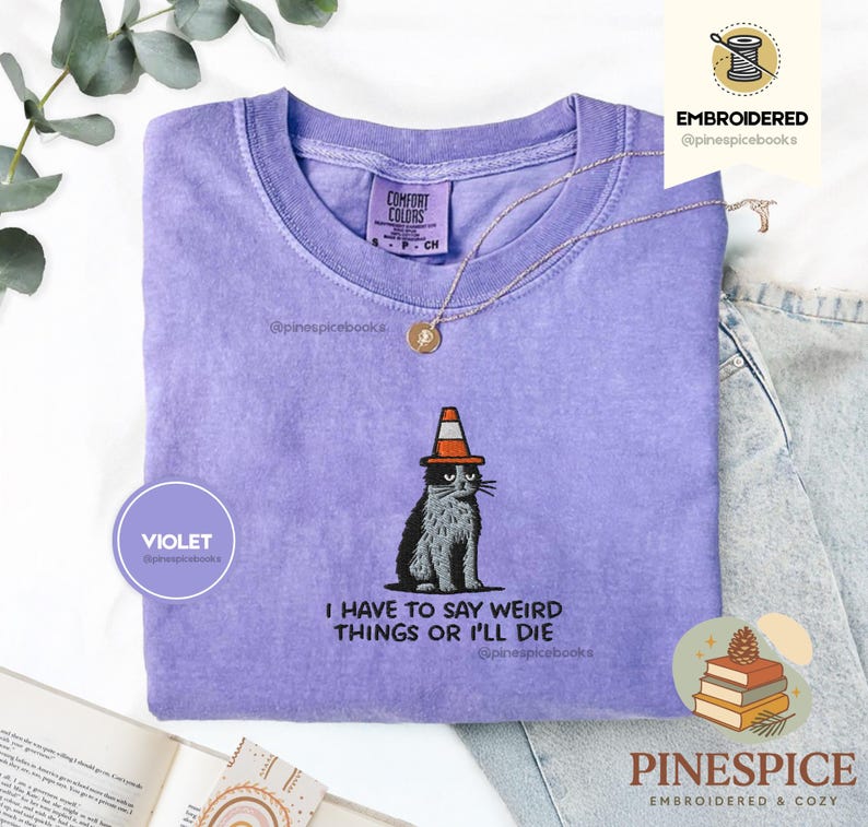 May include: A violet Comfort Colors t-shirt with an embroidered design of a cat wearing a traffic cone hat. The text "I HAVE TO SAY WEIRD THINGS OR I'LL DIE" is below the cat. A gold necklace is on the shirt.