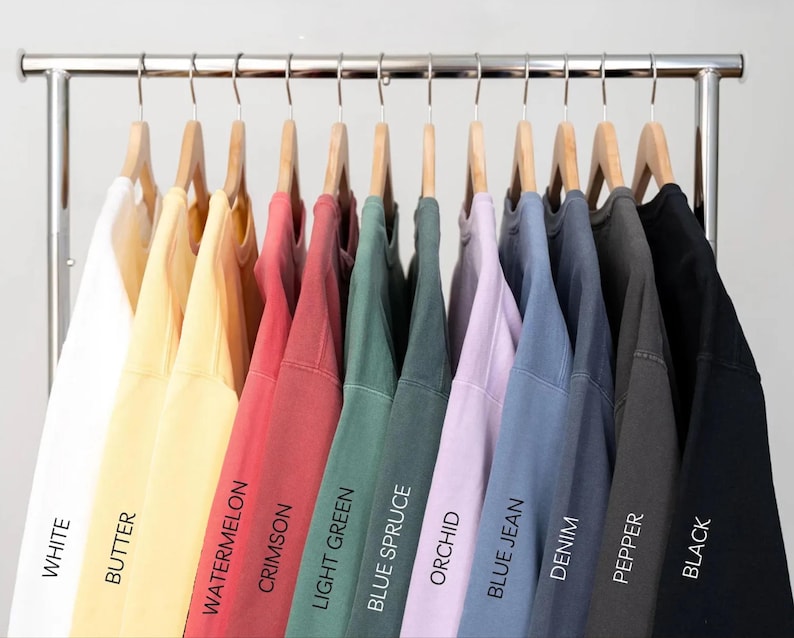 Puede incluir: Una fila de sudaderas de varios colores colgadas en perchas de madera. Los colores incluyen blanco, amarillo, rojo, verde, morado, azul, gris y negro. Cada sudadera tiene una etiqueta con el nombre de su color: Blanco, Mantequilla, Sand&iacute;a, Carmes&iacute;, Verde claro, Abeto azul, Orqu&iacute;dea, Blue Jean, Denim, Pimienta y Negro.