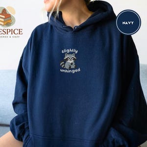 Pu&ograve; includere: Felpa con cappuccio blu navy con la scritta ricamata "slightly unhinged" e una grafica di un procione. La felpa ha un cappuccio con coulisse e una tasca a marsupio. Il marchio "Pinespice" &egrave; visibile in alto a sinistra.