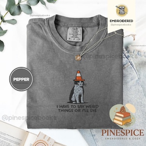 May include: A gray Comfort Colors t-shirt featuring an embroidered cat wearing a traffic cone hat. The text "I HAVE TO SAY WEIRD THINGS OR I'LL DIE" is below the cat. A silver necklace with a pendant is also visible.