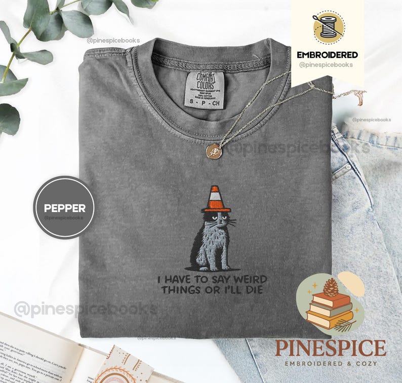 May include: A gray Comfort Colors t-shirt featuring an embroidered cat wearing a traffic cone hat. The text "I HAVE TO SAY WEIRD THINGS OR I'LL DIE" is below the cat. A silver necklace with a pendant is also visible.