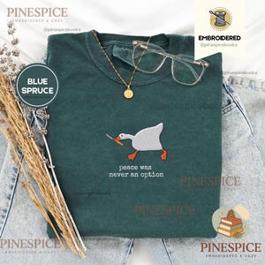 May include: A dark green t-shirt featuring an embroidered white goose holding a knife in its beak, with the text "peace was never an option" stitched below. The shirt is styled with a gold-tone necklace and clear-framed glasses.