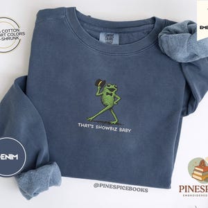 Puede incluir: Sudadera azul denim con un bordado de una rana verde con pajarita y sombrero de copa. El texto "THAT'S SHOWBIZ BABY" está debajo. Hecha de 100% algodón, pre-encogida y en colores confort.