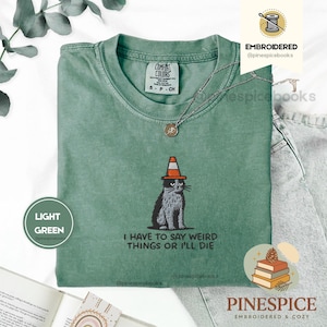 May include: Light green t-shirt with a graphic of a cat wearing a traffic cone hat. The text "I HAVE TO SAY WEIRD THINGS OR I'LL DIE" is below the cat. The shirt has a crew neck and short sleeves.